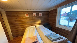 Ein blockhaushaltsähnliches Zimmer mit zwei Betten, Holzwänden, einem Fenster mit Aussicht und einem kleinen Nachttisch mit Lampe.