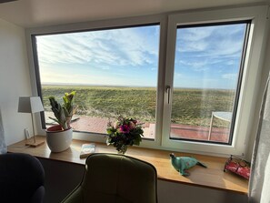 Een kamer met een groot raam, een bureau met een lamp, een stoel, een potplant, een vaas met bloemen, en een kleine tafel met een stoel erbuiten.
