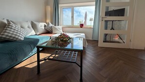 Une pièce comprenant un canapé, une table basse en verre agrémentée d’un arrangement floral, une fenêtre avec des rideaux et une porte dotée de panneaux de verre.