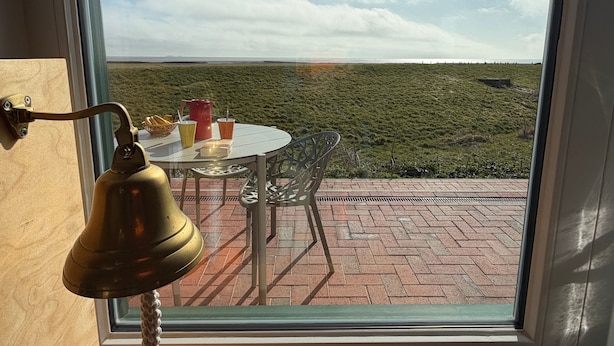 Une table avec une cloche, une bouilloire rouge et un panier de pain, le tout placé sur un balcon donnant sur un champ verdoyant.