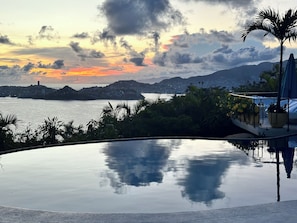 Ein Pool spiegelt einen Sonnenuntergang wider, mit Blick auf eine ferne Stadt und Berge.