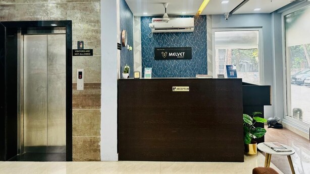 A reception desk with a sign reading 'MELVET,' an elevator, and a reception area with a window view. - HOTEL MELVET RESIDENCY (Kanayannur)
