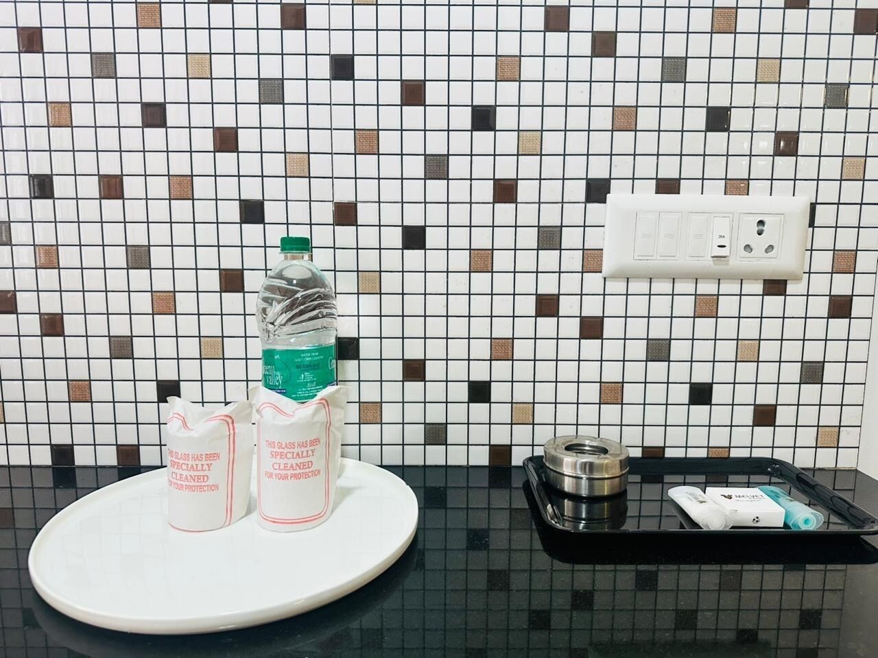 Een keukentafel met een wit bord, twee glazen bakjes, een fles, een dienblad met een metalen bakje, en een muur met een betegelde spatwand en stopcontacten.