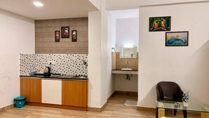 A modern kitchen with a tiled backsplash, wooden cabinets, a sink, and a countertop. There are framed artworks on the wall and a chair in the corner. - HOTEL MELVET RESIDENCY (Kanayannur)