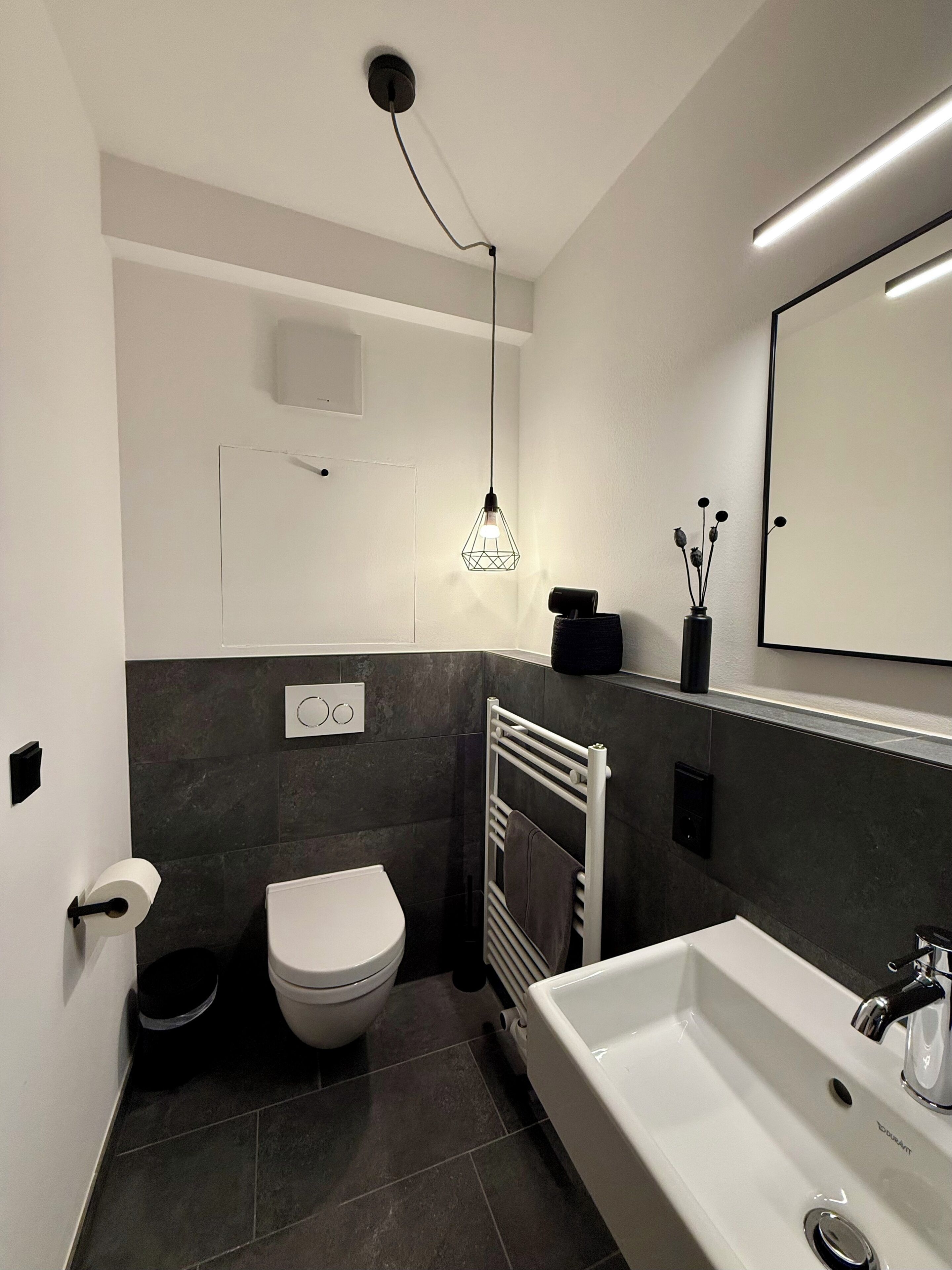 A modern bathroom with a white toilet, a white sink, a towel rack, a light fixture, and a mirror.