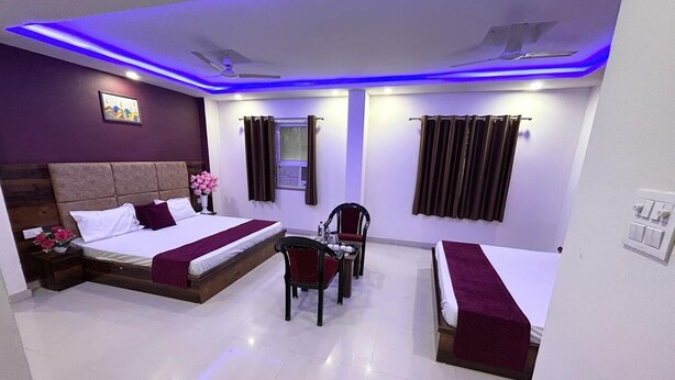 A hotel room with two beds, a desk, a chair, a small table, a window with curtains, and a ceiling fan. - Hotel Excotia Haridwar (Haridwar)