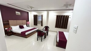 A hotel room with a bed, a desk, a chair, a window with curtains, and a ceiling fan. - Hotel Excotia Haridwar (Haridwar)