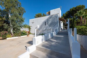 A white building with a staircase leading to a door, surrounded by greenery and a clear sky. - Villa Lantana with private pool - My Rental Homes (Santa Eulària Des Riu)
