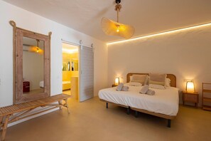 A bedroom with a bed, bedside lamps, a wooden bench, a mirror, and a door leading to a bathroom. - Villa Lantana with private pool - My Rental Homes (Santa Eulària Des Riu)