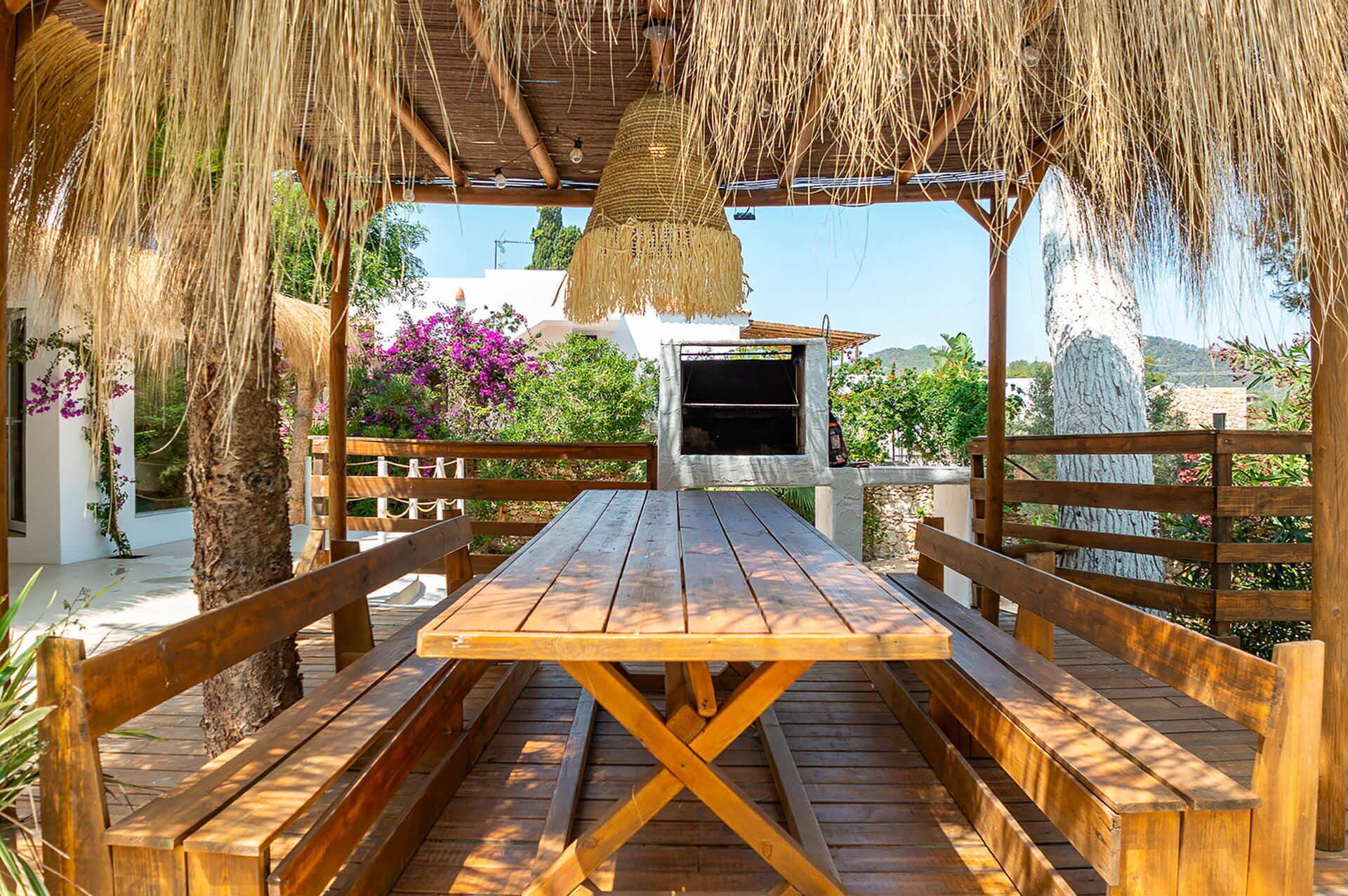 An outdoor wooden table with benches, a barbecue grill, and a thatched roof structure.