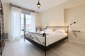 A bedroom with a metal bed frame, white bedding, a window with curtains, and a hanging light fixture. - Chuchotis, Apartment for 4 with a balcony and parking space (LE Crotoy)