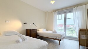 A hotel room with two beds, a wooden nightstand, a small cabinet, and a large window with curtains. - Chuchotis, apartment for 4 with balcony and parking space (LE Crotoy)