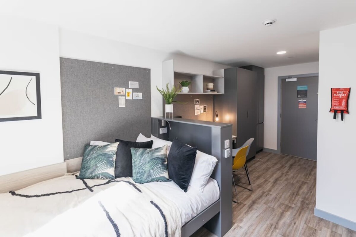 A modern bedroom with a bed, a desk, and a chair. There is a small kitchenette area with a fridge and a sink.