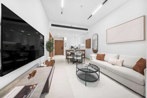 A modern living room with a large flat-screen TV, a dining area with a table and chairs, a sofa with cushions, and a large abstract painting on the wall. - StayShort - Charming 1BR Retreat in JVC Sleeps 4 (JVC)