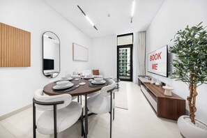 A modern dining area with a round table, white chairs, a wooden panel, a mirror, a TV, and a potted plant. - StayShort - Charming 1BR Retreat in JVC Sleeps 4 (JVC)