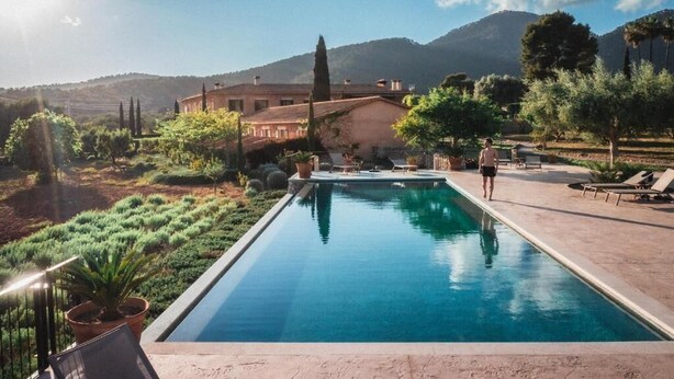 A rectangular swimming pool with a clear blue water surface, surrounded by a landscaped garden with various plants and trees, and a large building in the background. - Finca Es Cabàs (Santa Maria del Camí)