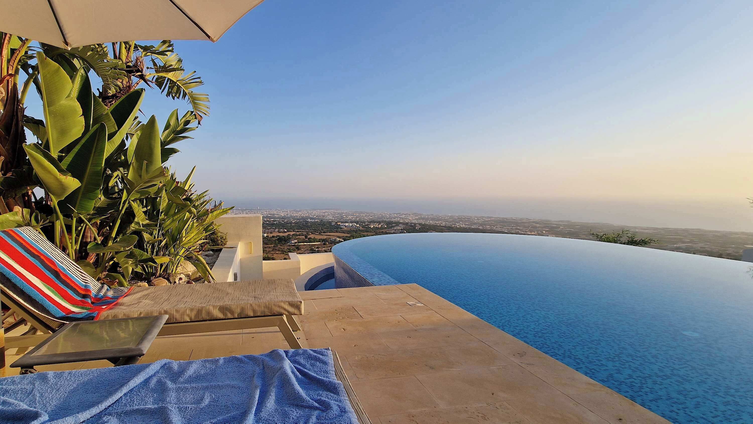 A infinity pool with a view of a cityscape, a lounge chair, and tropical plants.