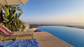Une piscine à débordement offrant une vue sur le paysage urbain, un fauteuil de relaxation et des plantes tropicales.
