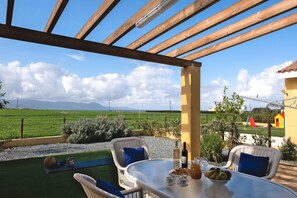 A patio with wicker chairs, a table with food and drink, and a view of a green field and mountains. - Special Launch Price! Amazing Sea View 1BR Villa, Garden (Polis Chrysochous)