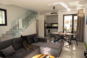 Ein modernes Wohnzimmer mit grauem Sofa, einer Glasstiege, einem Essbereich mit Tisch und Stühlen und einer Küche im Hintergrund.