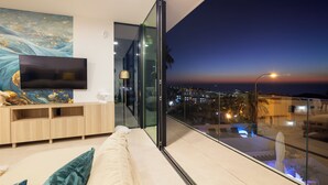 A modern living room with a flat-screen TV, a sofa, and a glass door leading to a balcony with a city view at dusk. - 200m NEW A Villa with private, heated pool and amazing ocean view (Costa Adeje)