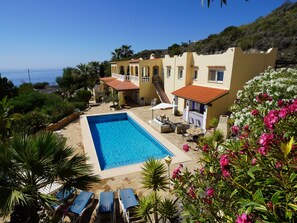 Ein Poolbereich mit freiem Blick auf das Meer, ein Gebäude mit rotem Ziegeldach und eine Treppe, die zu einer oberen Ebene führt.