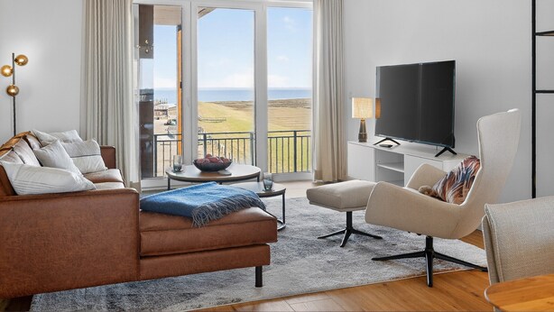 Ett modernt vardagsrum med en soffa, fåtölj, TV och utsikt över landskapet genom stora fönster.