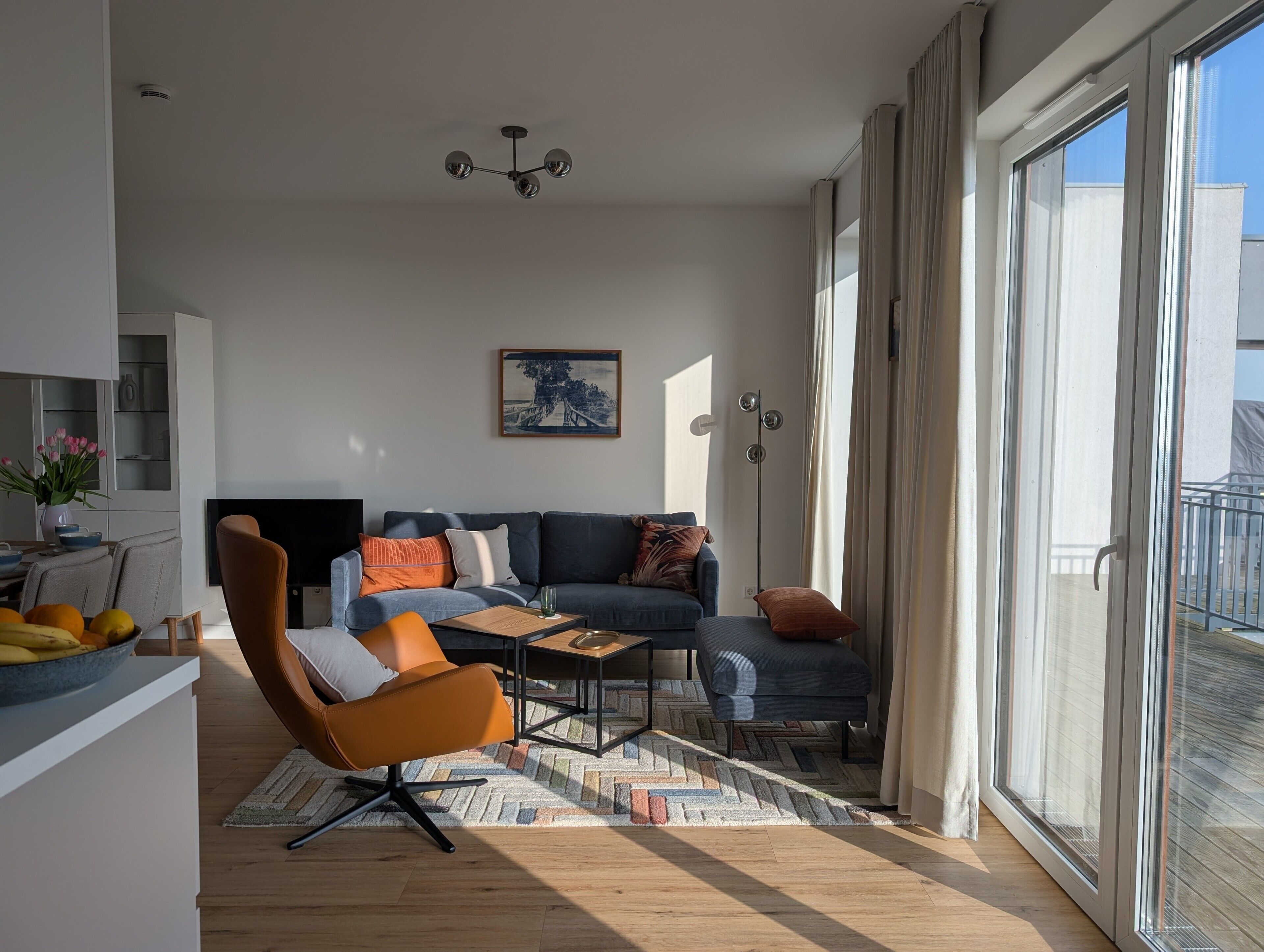 A modern living room with a sofa, coffee table, and a chair. There is a painting on the wall, a ceiling light, and a window with curtains.