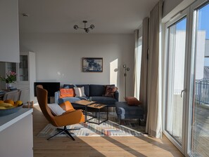 A modern living room with a sofa, coffee table, and a chair. There is a painting on the wall, a ceiling light, and a window with curtains. - Penthouse 'Smilla' with Sea View and Wi-Fi (Pellworm)