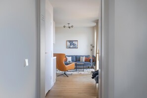 Un pasillo que conduce a una sala de estar con un sofá azul, un sillón naranja y una lámpara en el techo.