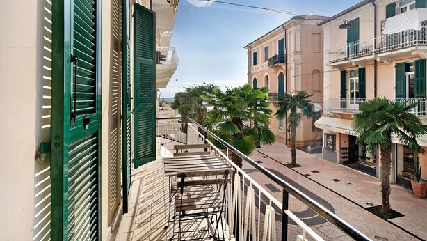 Een balkon met een metalen hek, een straat met palmbomen en gebouwen met balkons en luiken.