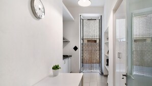 Een smalle gang met een witte tafel en stoelen, een klok aan de muur, en een deur naar een kamer met een kamerbehang.'