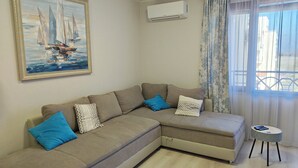 A living room with a beige sectional sofa, a painting of sailboats, an air conditioning unit, and a window with curtains. - Ines - Golf and the Sea in Bulgaria (Balchik)