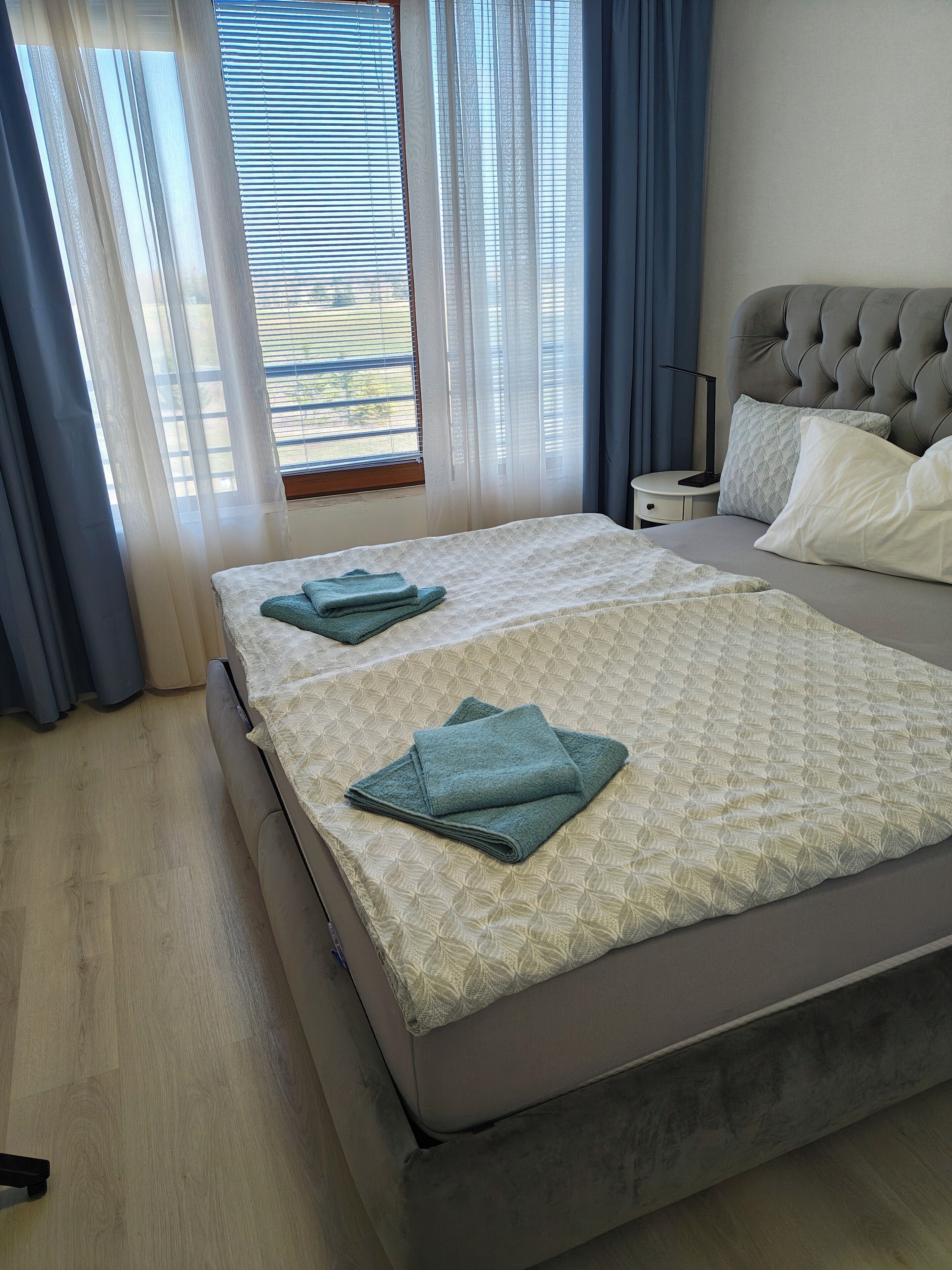 A neatly made bed with a patterned bedspread, a bedside table with a lamp, and a view through a window with blinds.