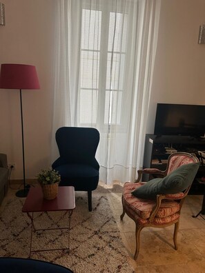 A living room with a dark blue armchair, a red floor lamp, a patterned rug, a wooden chair with a cushion, and a television. - Apartment & TerraceS t-Rémy Conciergerie Maurin (SAINT REMY DE PROVENCE)