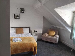 Een slaapkamer met een bed, een stoel, een nachtkastje en een raam met een wit plafond.