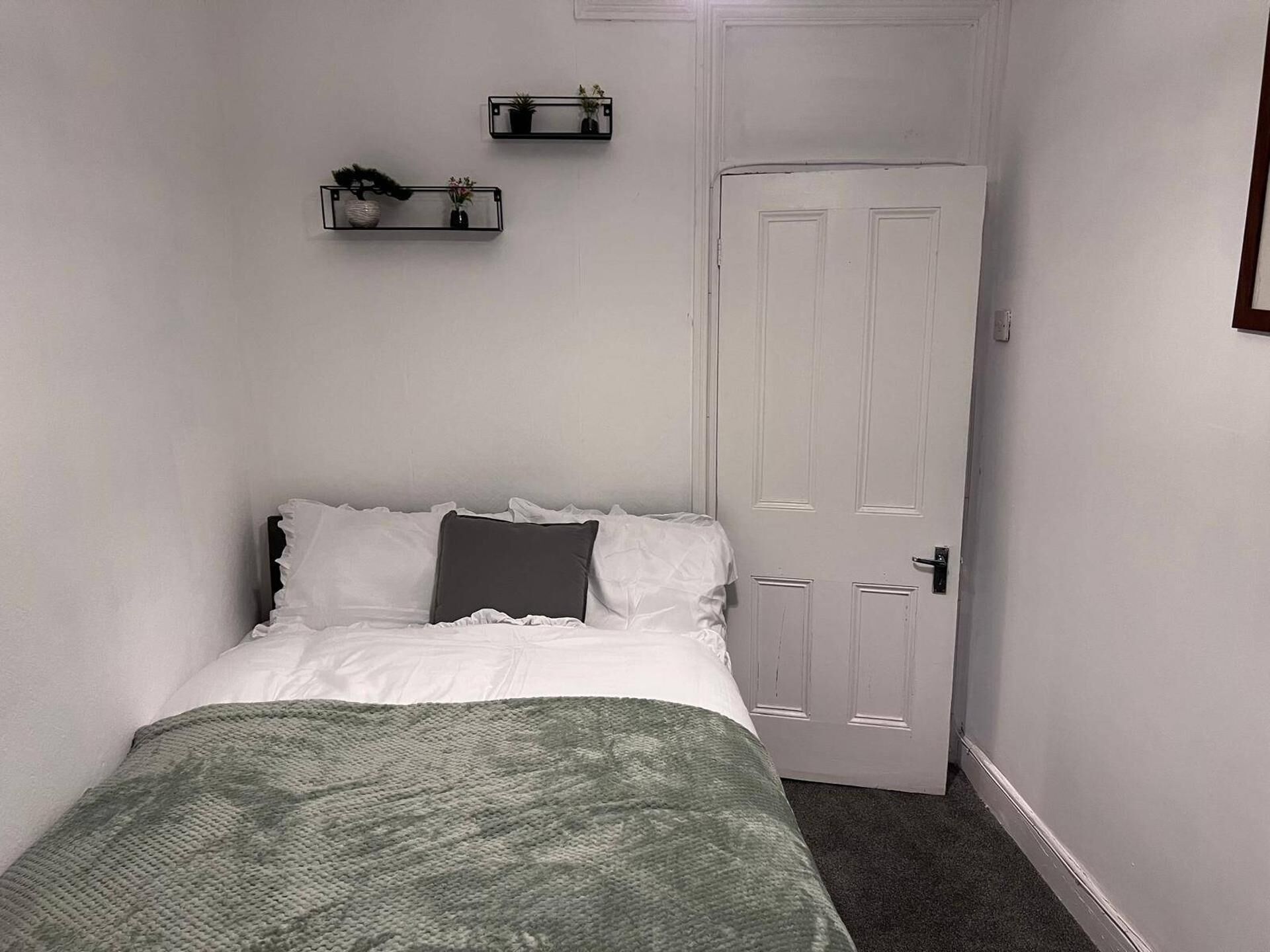 Una habitación pequeña y estrecha con una cama individual, una puerta blanca y dos estantes en la pared con plantas en macetas.
