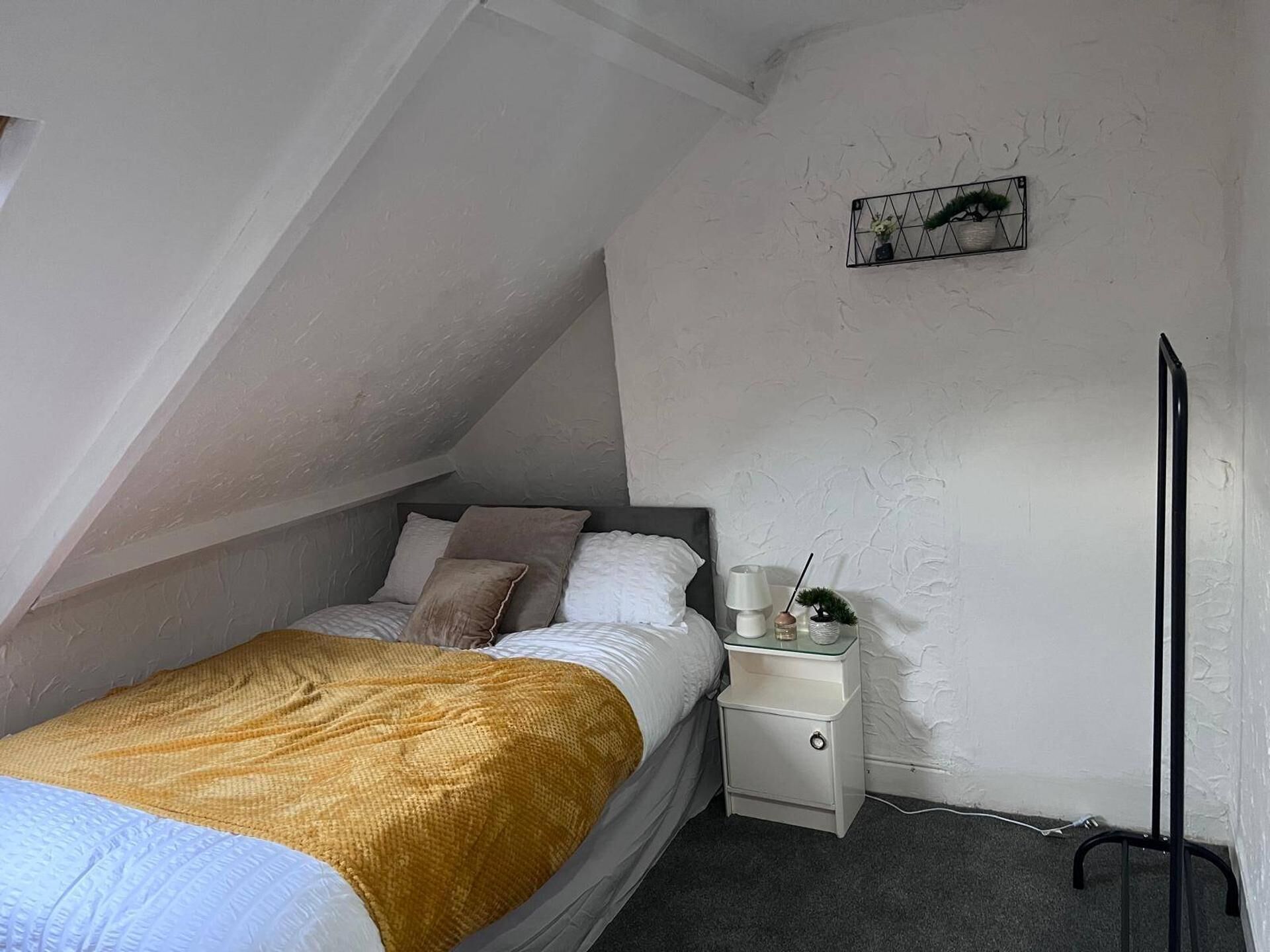 Une chambre avec un plafond en pente, un lit recouvert d’une couverture jaune, une table de chevet avec une lampe et un tableau accroché au mur.