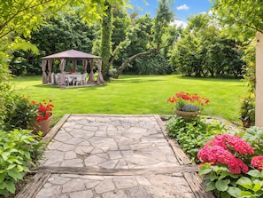 A stone patio leading to a gazebo with a table and chairs, surrounded by lush greenery and flowering plants.