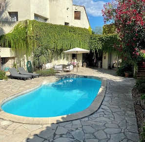 A swimming pool with stone tiles, a white umbrella, and a white building with green vines.