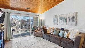 Una sala de estar moderna con techo de madera, un sofá seccional grande y un ventanal corredizo que da a un balcón.