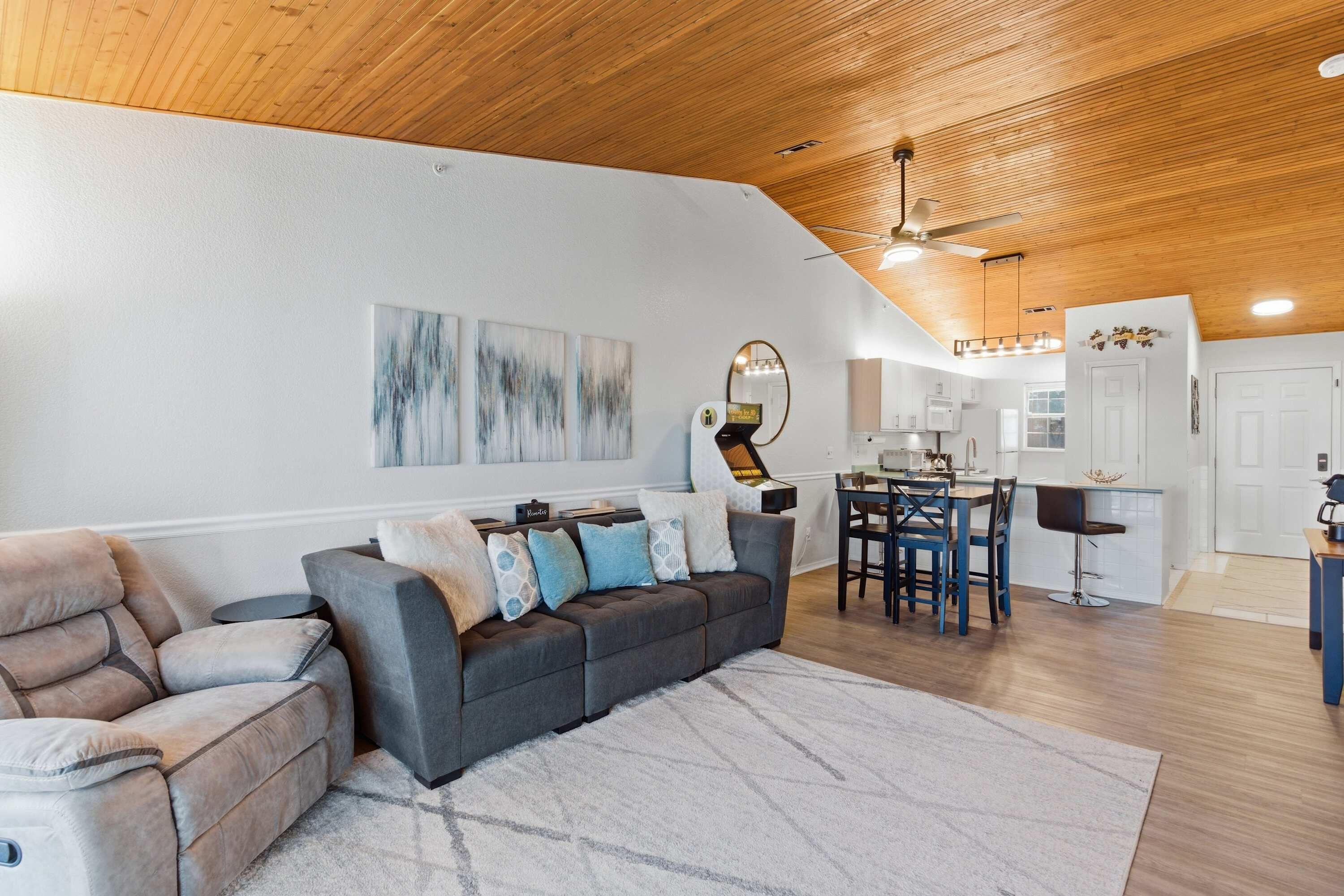 A modern living room with a sectional sofa, a dining area with a table and chairs, a kitchen with white cabinets, and a ceiling fan.