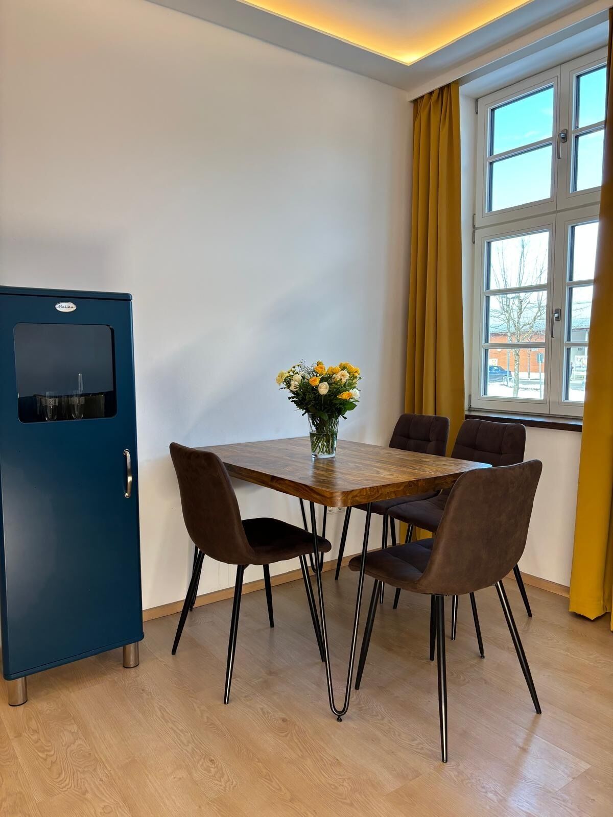A room with a wooden table, four chairs, a blue cabinet, and a window with yellow curtains.