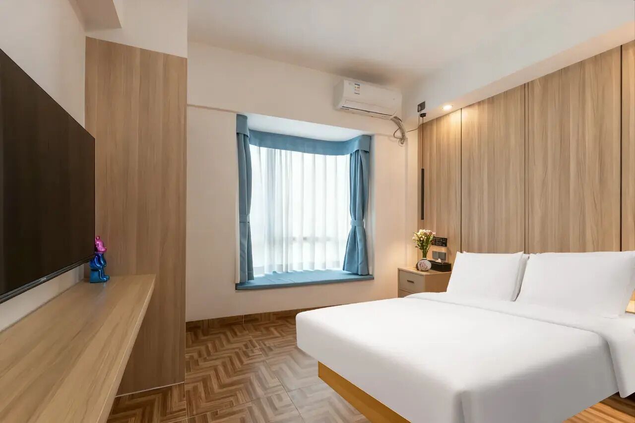 Ein Hotelzimmer mit Bett, Fernseher, Fenster mit Vorhängen, einer Wand-Klimaanlage und Holzboden.