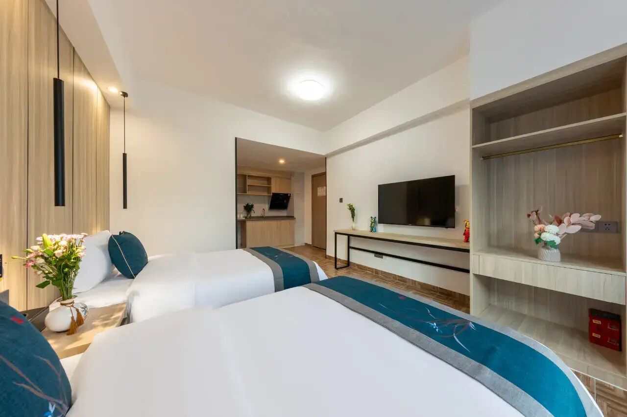 A hotel room with two beds, a TV, a small kitchenette, and a TV stand with a flower arrangement.