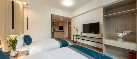 A hotel room with two beds, a TV, a small kitchenette, and a TV stand with a flower arrangement.