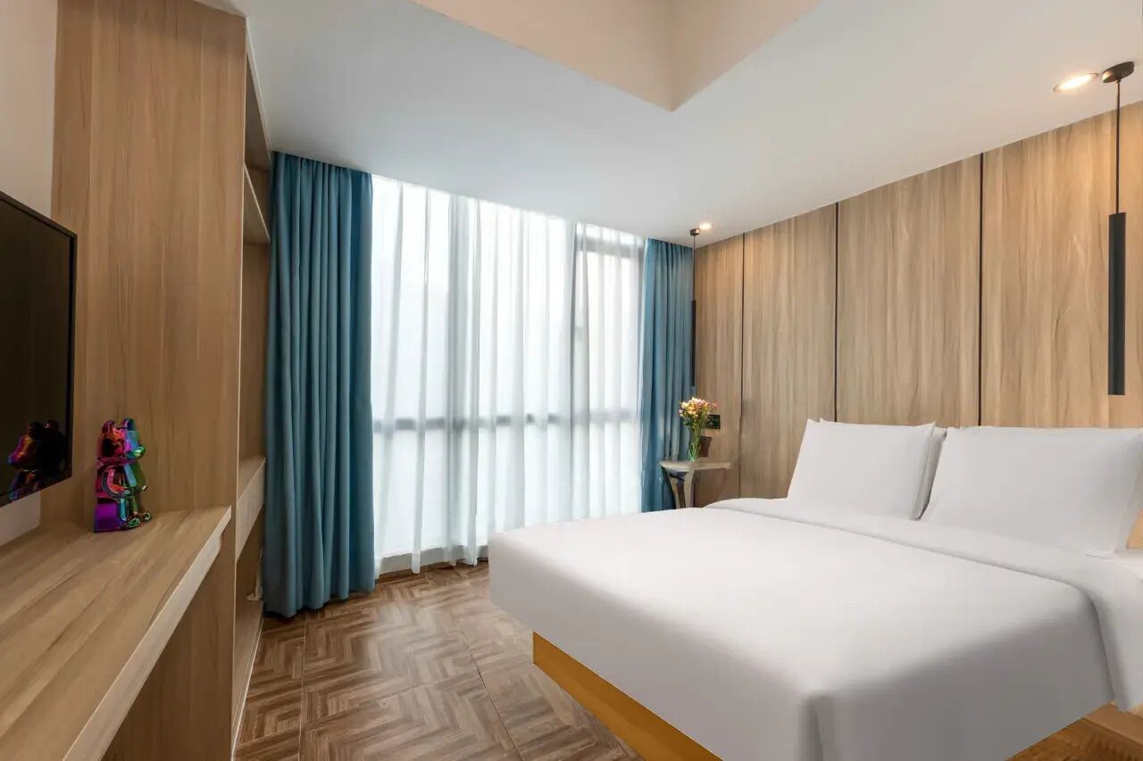 Une chambre d’hôtel avec un lit, une télévision, une petite table avec un vase et une fenêtre avec des rideaux.
