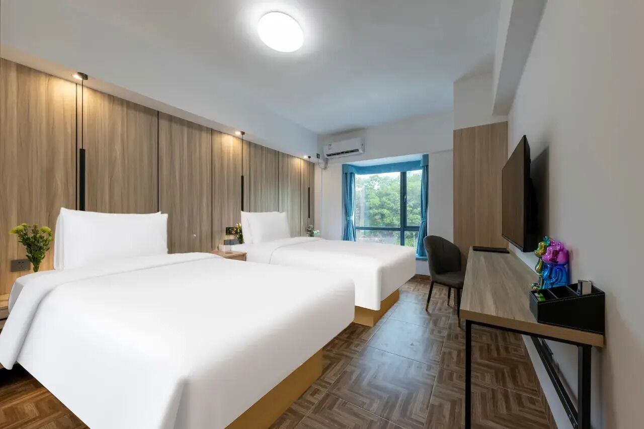 Ett hotellrum med två sängar, ett skrivbord, en stol, en TV och ett fönster med utsikt över grönområden.