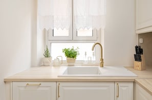 Eine Küche mit weißem Spülbecken, einem Topf mit Pflanze, einem Fenster mit Spitzenvorhängen und einem Messerblock.