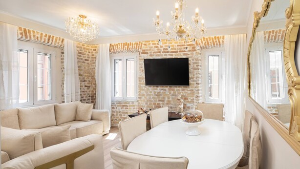 A dining area with a brick wall, a flat-screen TV, a chandelier, and a round table with chairs. - Borgo Veneziano by Corfuescapes (Kerkira)